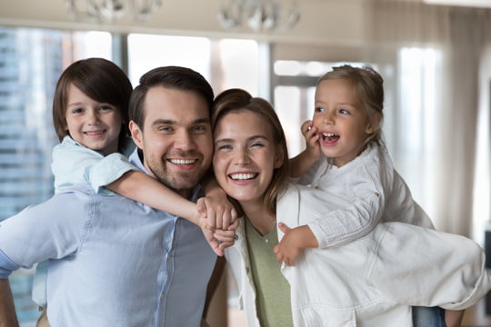 Cheerful young parents piggy backing little kids, holding children Cheerful young parents piggy backing little kids, holding children, looking at camera with toothy smile, promoting white healthy teeth, dental care, dentistry service. Family Dentistry